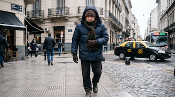 Un hombre abrigado con campera azul, bufanda y gorro camina por una calle de adoquines junto a un taxi y un autobús.