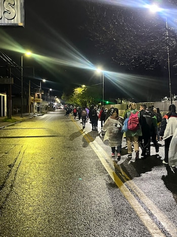 Varias personas vistas de espaldas caminan de noche por una calle asfaltada y mojada iluminada por farolas