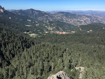 Vista aérea de montañas cubiertas por densos bosques, un valle con un poblado de casas y tejados rojos, cielo azul claro y rocas en primer plano