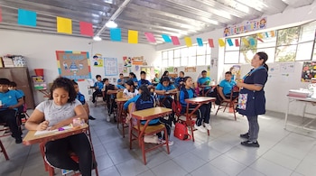 Una docente de pie imparte clase a varios alumnos sentados en pupitres dentro de un aula decorada con guirnaldas de colores