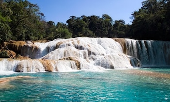 Una vista panorámica de una cascada de agua sobre rocas escalonadas, con agua de tono turquesa en la base y vegetación frondosa en los laterales bajo un cielo azul