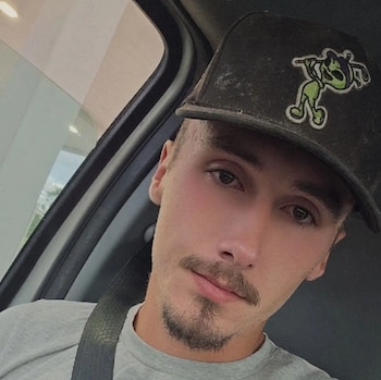 Un hombre joven con bigote y gorra negra mira a la cámara sentado en el interior de un automóvil con el cinturón puesto