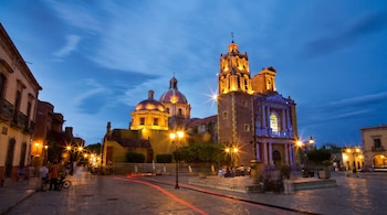 Vista nocturna de una plaza con un templo colonial iluminado, cúpulas, campanario, farolas, personas, edificios antiguos y rastros de luces de vehículos.