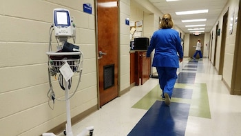 Pasillo de hospital. Una enfermera en uniforme azul camina de espaldas junto a un monitor de signos vitales. El suelo tiene un patrón geométrico azul, blanco y verde