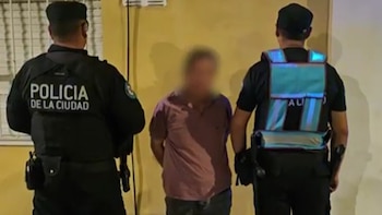 Vista trasera de dos oficiales de la Policía de la Ciudad escoltando a una persona borrosa en el centro, contra una pared de color claro. Los oficiales visten uniformes oscuros