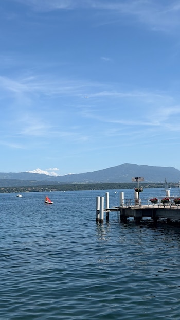 Un lago azul bajo un cielo despejado, con montañas y picos nevados a lo lejos; un muelle con macetas florales y una balsa inflable con tobogán en el agua