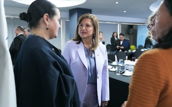 Una mujer con traje formal de color lila conversa de frente con dos personas en una sala con mesas y más asistentes
