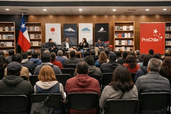 Auditorio con personas sentadas de espaldas, cuatro ponentes en un escenario, estanterías con libros, bandera chilena y cartel rojo de ProChile.