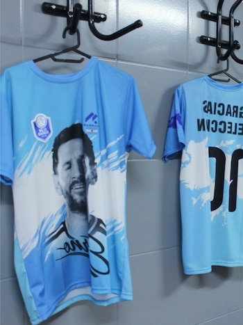 Dos camisetas de fútbol celestes cuelgan de ganchos en una pared de azulejos grises. Una exhibe la imagen de Lionel Messi y la otra, el número diez