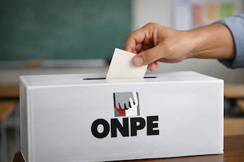 Primer plano de una mano introduciendo una papeleta en una urna electoral blanca con el logo de la ONPE, sobre un fondo borroso de aula escolar.