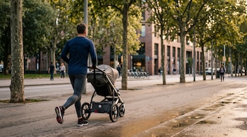 Vista posterior de un hombre con ropa deportiva que corre y empuja un cochecito de bebé por una acera con árboles y edificios.