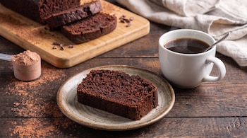 Rebanada de panqué de chocolate en plato, taza de café con cuchara, panqué rebanado en tabla, pala medidora con polvo de cacao y trozos de cacao en mesa de madera.