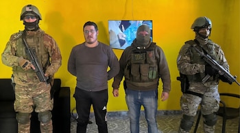 Cuatro hombres en una habitación con paredes amarillas. Dos militares armados flanquean a un hombre en ropa civil y a otro con balaclava y chaleco táctico.
