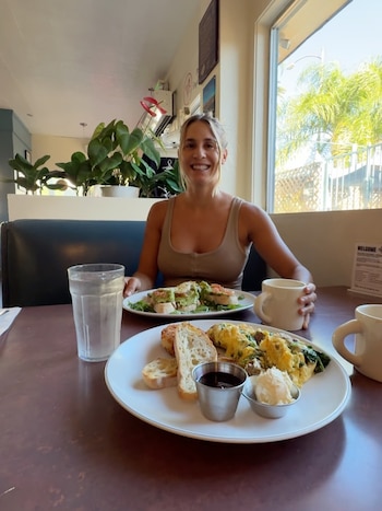 Mujer rubia sonriente sentada en una mesa de restaurante con dos platos de comida, un vaso de agua y tazas de café, frente a una ventana soleada