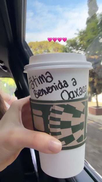 Una mano sostiene una taza de café con mensaje 'Fátima Bienvenida a Oaxaca' y protector de cartón. Cinco corazones rosas flotan sobre la taza. Cielo azul y árboles