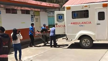 Varias personas trasladan a un paciente en camilla hacia una ambulancia blanca con las puertas traseras abiertas