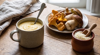 Taza con polenta, cuchara, plato con hongos chanterelles y porcini, pote de queso rallado con cuchara de madera, servilleta, mesa de madera, ventana.
