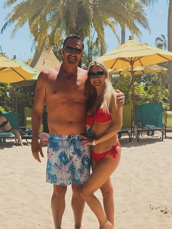 Un hombre y una mujer sonrientes posan en una playa soleada. El hombre lleva bañador y la mujer un bikini rojo, con palmeras y sombrillas de fondo