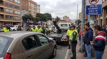 Imagen de una calle en Bogotá donde un coche gris con la ventana rota está siendo remolcado por una grúa, rodeado por policías y civiles.