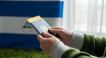 Primer plano de manos adultas sosteniendo un pasaporte azul oscuro con un sello dorado. Al fondo, una bandera de Nicaragua borrosa y luz fría de una ventana.