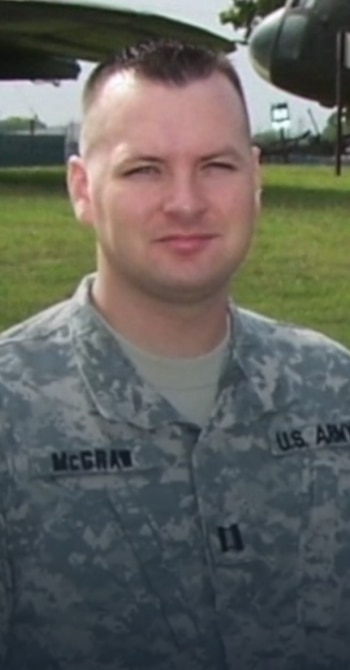 Hombre con uniforme militar de camuflaje, placa de identificación con el apellido McGraw, y el texto US ARMY en el hombro. Fondo borroso con vegetación y helicóptero