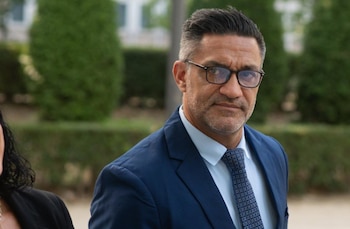 Hombre con traje azul, camisa celeste y corbata estampada, con gafas. Fondo verde borroso con vegetación. Parte del hombro y pelo de otra persona