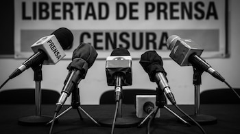 Imagen en blanco y negro de cinco micrófonos de conferencia sobre una mesa, algunos cubiertos con tela negra, frente a un cartel que dice "LIBERTAD DE PRENSA CENSURA".