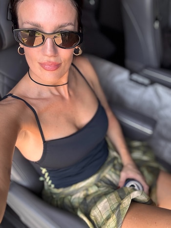 Mujer con gafas de sol oscuras, top negro de tirantes, falda a cuadros verde, aros dorados y choker negro. Está sentada en un automóvil
