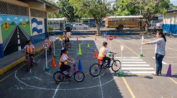 Niños en bicicletas con chalecos de seguridad practican normas de tránsito en un patio escolar con conos y señales, guiados por una mujer.