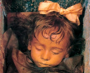 Primer plano de la cabeza de una momia infantil con cabello castaño, ojos cerrados y un lazo color crema en la parte superior de la cabeza