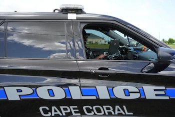 Vehículo patrullero negro de la Policía de Cape Coral con distintivos azules. Un oficial con gafas de sol trabaja en una laptop dentro del automóvil