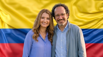 Retrato de una mujer y un hombre sonriendo a la cámara, con la bandera de Colombia, de franjas amarillas, azules y rojas, como fondo.