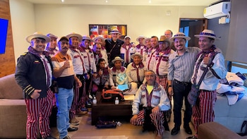 Un grupo de más de quince hombres con sombreros y pantalones de rayas posa de pie y sentados en una sala junto a una mesa de centro