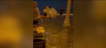 Cuatro personas están de pie en medio de agua acumulada junto a una columna y una estructura metálica por la noche