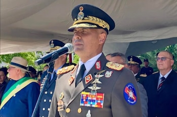 Primer plano de un hombre con uniforme militar y gorra, hablando ante un micrófono, con condecoraciones en el pecho, y otras personas detrás