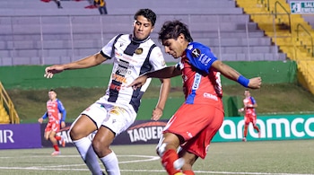 Dos jugadores de fútbol vestidos con uniformes, uno blanco y negro, el otro rojo y azul, en un campo de césped con líneas blancas, bajo luces de estadio.