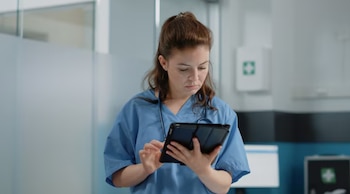 Una mujer con cabello castaño recogido, vestida con uniforme médico azul, usa una tableta con ambas manos. Al fondo, un botiquín de primeros auxilios y paredes blancas