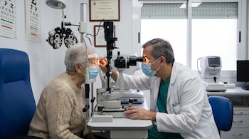 Un médico examina el ojo de una paciente mayor con una lámpara de hendidura en un consultorio oftalmológico; ambos usan mascarillas.