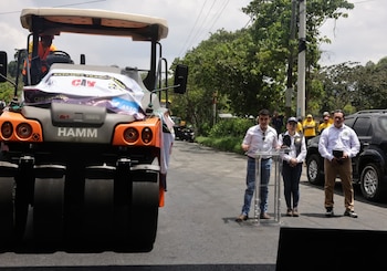 Vista trasera de un rodillo de asfalto gris y naranja de la marca HAMM con un cartel blanco, y tres personas frente a un podio en una carretera pavimentada