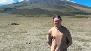 Hombre con bigote y gafas de sol en la cabeza, viste camiseta clara, posa de pie en un campo de pasto seco con una montaña nevada al fondo bajo cielo azul