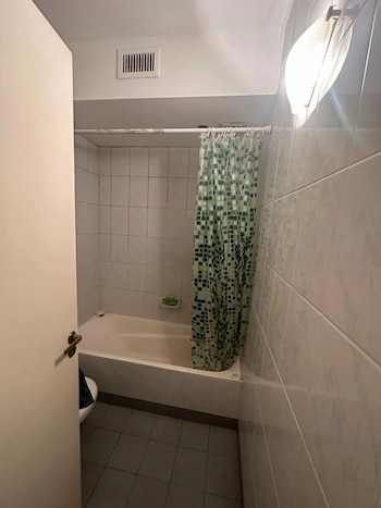 Vista interior de un baño con azulejos blancos, una bañera, cortina de ducha verde y blanca, y un suelo de baldosas grises