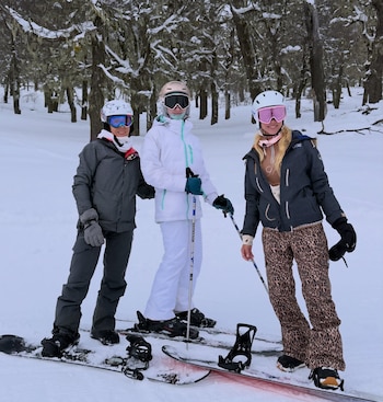 Tres personas con cascos, antiparras, chaquetas, guantes. Una sobre snowboard, otra sobre esquís. Dos sujetan bastones de esquí. Nieve y árboles