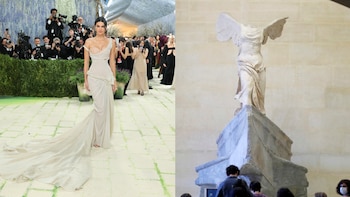 Kendall Jenner con un vestido beige claro de hombros caídos y larga cola en una alfombra verde, junto a la estatua de mármol sin cabeza de la Victoria Alada de Samotracia con alas