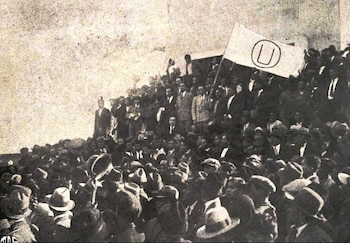 Fotografía antigua en blanco y negro de una multitud de hombres con trajes y sombreros, algunos sosteniendo una bandera blanca con la 'U' de Universitario