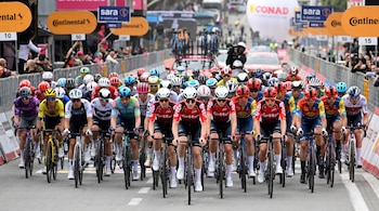 Vista frontal de un gran grupo de ciclistas con cascos y uniformes de varios colores, pedaleando en una carrera bajo pancartas de patrocinadores.