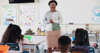 Una profesora sonriente de pie frente a una pizarra en un aula, mientras varios estudiantes la observan sentados en sus pupitres