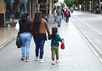 Tres personas, dos adultos y una niña, caminan por una acera gris. La niña lleva una mochila roja y está de la mano de un adulto. Edificios y una calle a un lado