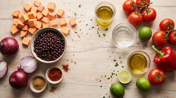 Batata en cubos, frijoles negros, cebollas, tomates, pimiento rojo, limas, especias en polvo, recipientes con aceite y agua sobre madera.
