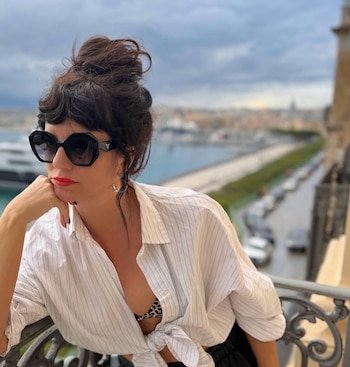 Mujer con gafas de sol oscuras y cabello recogido en un moño alto, luciendo una camisa desabrochada sobre un top de animal print, en un balcón con vista a un puerto y una ciudad