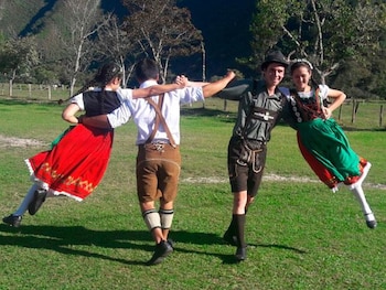 Cuatro personas vestidas con trajes tradicionales austro-alemanes, dos hombres y dos mujeres, posan o danzan en un campo de hierba verde con colinas y árboles al fondo